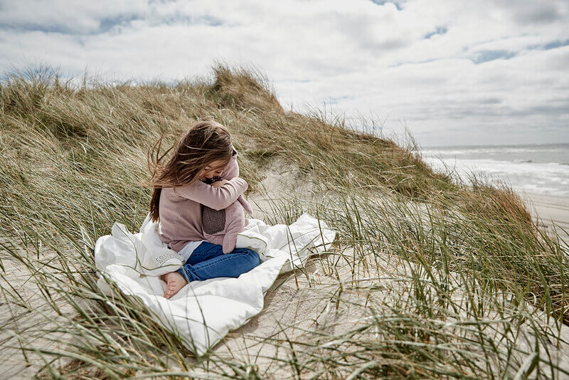 Girl sitting on a Cocoon Amazing Maize duvet hugging a teddy bear on a beach in Denmark